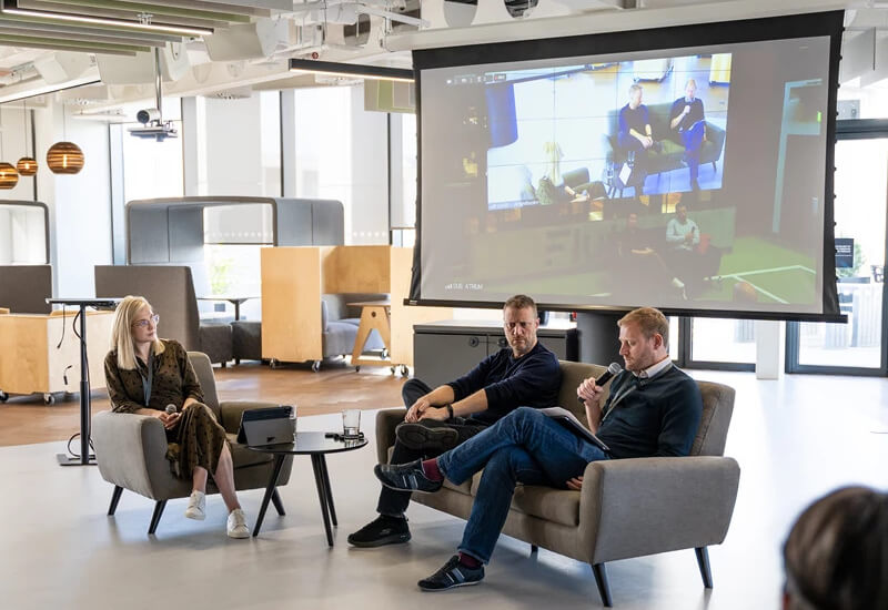 Three colleagues engaged in a discussion while a projector screen shows content in the background