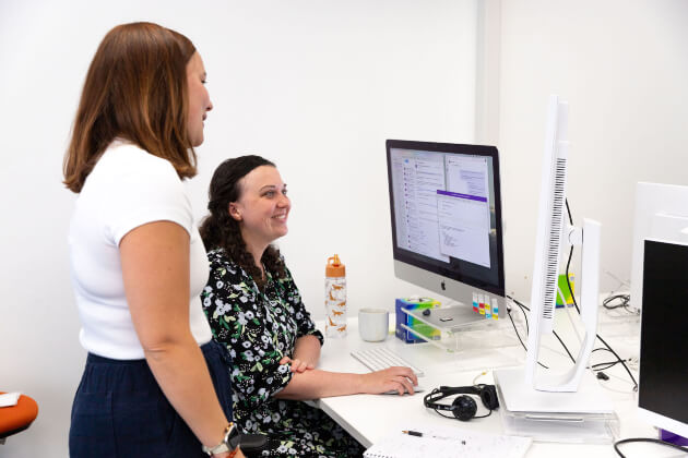 One female sitting and one female staying in front of a monitor