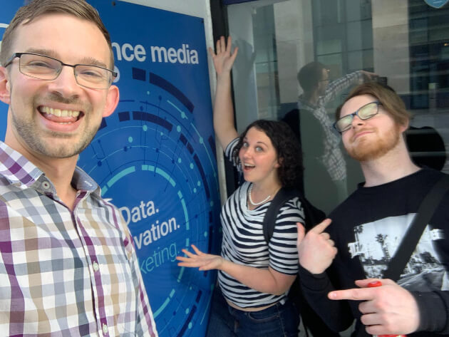 Three colleagues in front of the Enhance Media banner